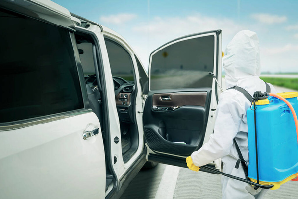 Male worker wearing protective suit while spraying disinfectant to car interior on the roadside for preventing coronavirus transmission - Fotoğraf, Görsel