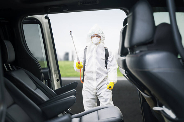 Young man wearing protective suit while spraying disinfectant to car interior on the roadside for preventing coronavirus transmission - Photo, image