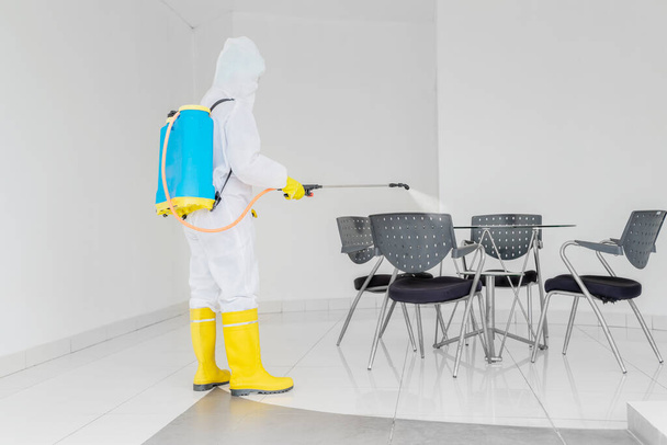 Young man wearing protective suit while spraying disinfectant to table and chairs in house during coronavirus pandemic - Фото, изображение