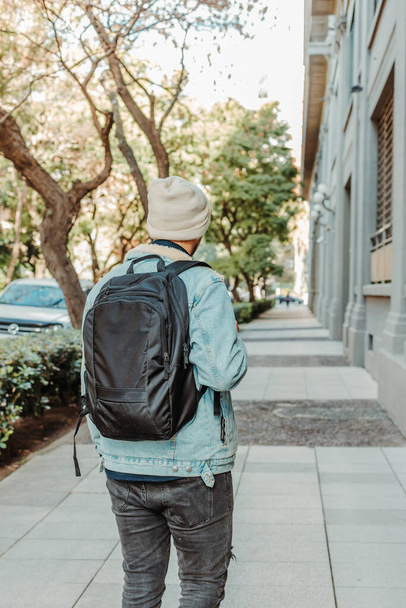 A back view of a young stylish male walking on the street wearing a denim jacket and a black backpack - Foto, afbeelding