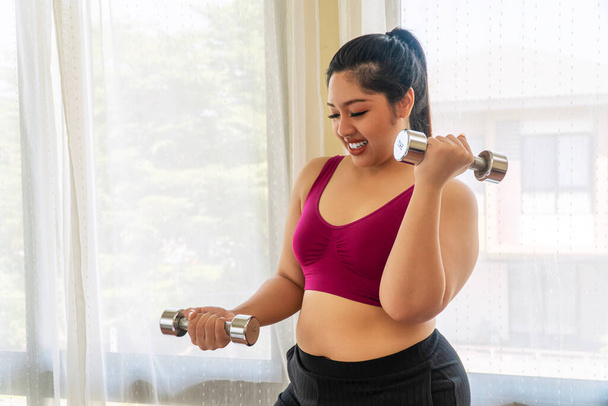 Άσκηση δραστηριότητας κατάλληλο σώμα για δίαιτα ή παχυσαρκία έννοια. Θετική ευτυχισμένη υπέρβαρη χοντρή γυναίκα κρατώντας αλτήρες στα χέρια προπόνηση δίαιτα απώλεια βάρους, ενώ στέκεται στο δωμάτιο. - Φωτογραφία, εικόνα