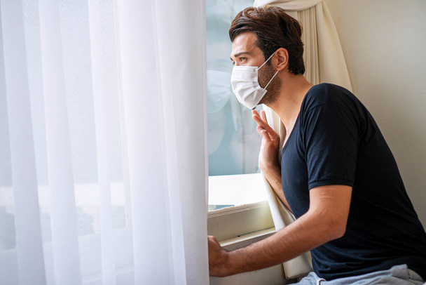 Outbreak Quarantine at home concept. Unhappy young man wear casual cloth and protective face mask worried something while sitting beside windows alone in the room.  - Foto, Bild