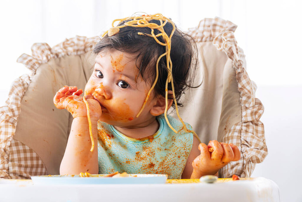 Adorable niña divertida comiendo espaguetis con cuchara mientras está sentada en una silla de alta potencia en casa. Niño pequeño con salsa de tomate haciendo lío su cara mirando a los padres. Concepto de autoalimentación - Foto, Imagen