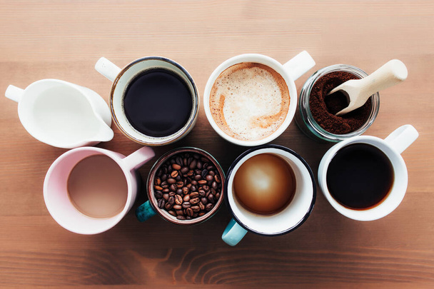 Liczne filiżanki kawy i wielobarwne kubki z americano, espresso, latte i cappuccino, słoik mleka, palone ziarna kawy w butelce szklanej, drewniana łyżka i mielona kawa w słoiku na drewnianym tle - Zdjęcie, obraz
