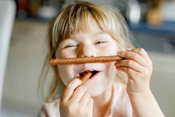 チョコレートワッフルロールを持つ幸せな小さな就学前の女の子の肖像画。甘いビスケットウェーハで空腹の幼児の子供を笑顔。甘い砂糖ココアワッフル. - 写真・画像
