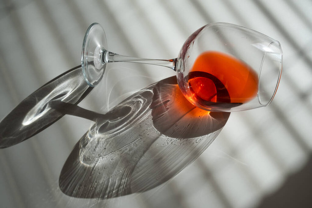 A glass of red wine lies on its side on a white table with a shade from the blinds - 写真・画像