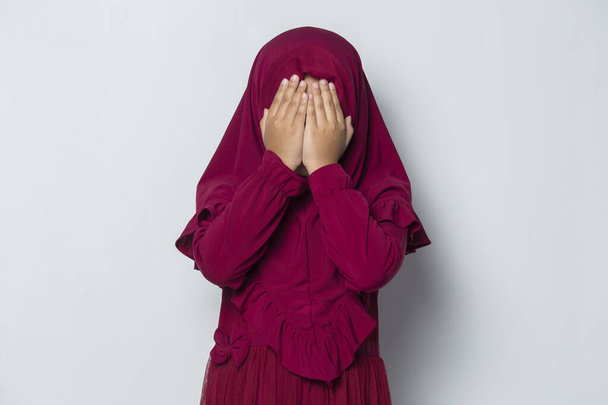 Φοβάται ασιατικό μουσουλμανικό κοριτσάκι καλύπτει το πρόσωπό της με τα χέρια της σε λευκό φόντο - Φωτογραφία, εικόνα