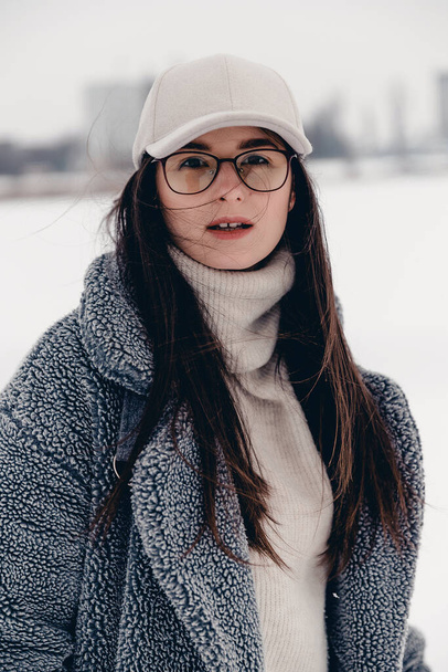 Portrait of an attractive girl dressed in stylish winter clothes. Woman posing for a photo on a snow-covered and icy lake in a local park - Foto, Imagem