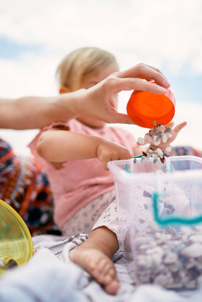La mano de la mujer vierte pequeñas piedras en un cubo de plástico al lado del niño. Primer plano - Foto, imagen