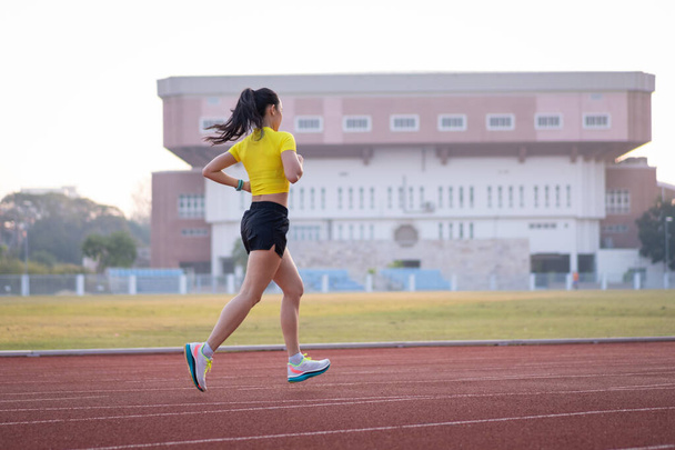 Genç bir Asyalı bayan atlet, formda ve sağlıklı yaşam tarzını korumak için güneşli bir sabahta şehir stadyumunda koşu pistinde koşuyor. Genç fitness kadını stadyum pistinde koşuyor. Spor ve eğlence. - Fotoğraf, Görsel