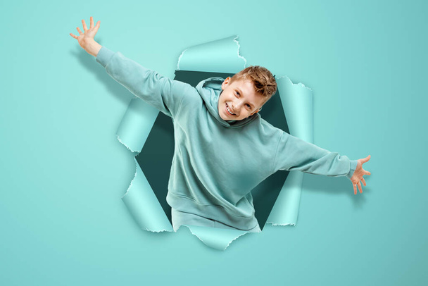 Boy on a blue background, happy waving his hands, punching a paper wall - 写真・画像