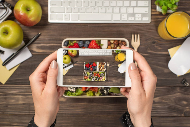 First person top view photo of hands holding smartphone taking photo of lunchboxes with healthy food glass of juice plant stationery keyboard mouse isolated dark wooden desk background with copyspace - Photo, Image