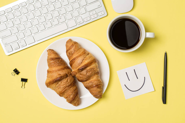 Top view photo of workplace white keyboard mouse binder clips pen sticker note with drawn smiling face cup of coffee and plate with two fresh croissants on isolated pastel yellow background - Foto, afbeelding