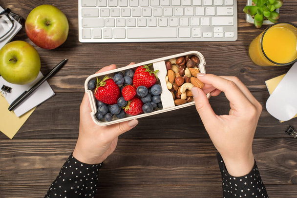 First person top view photo of woman's hands holding lunchbox with healthy meal nuts and berries over apples glass of juice flowerpot stationery keyboard mouse on isolated dark wooden table background - Foto, Bild