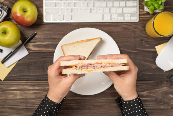 Фото от первого лица с женскими руками, держащими сэндвич над пластиной клавиатуры мышь яблоки стакан сока цветочный горшок и канцелярские принадлежности на изолированном темном деревянном столе фоне - Фото, изображение