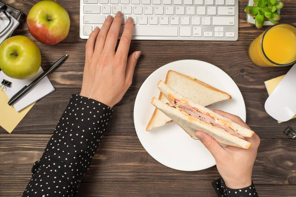 First person top view photo of female hands holding sandwich over plate and typing on keyboard mouse apples glass of juice flowerpot and stationery on isolated dark wooden table background - Photo, image