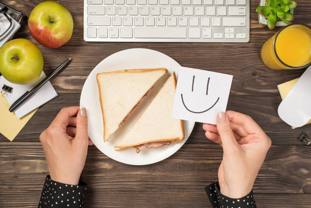 First person top view photo of hand holding sticker note with drawn smiling face over plate with toasts keyboard mouse apples glass of juice plant and stationery isolated dark wooden desk background - Фото, зображення