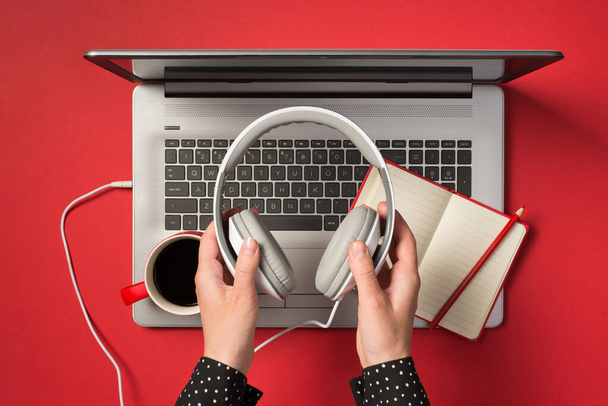 Oben Foto von grauen Laptop-Tasse Kaffee Notizbuch mit Bleistift und Händen hält einen Kopfhörer isoliert auf dem roten Hintergrund - Foto, Bild