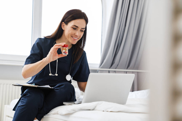 The smiling brunette woman doctor sitting on bed talking by video call and showing pills in jar to laptop camera in room - Φωτογραφία, εικόνα