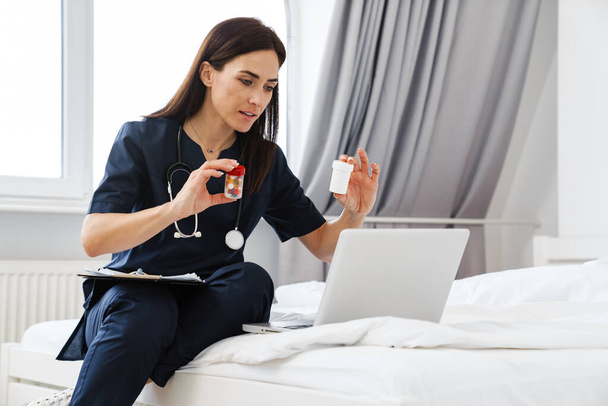 The brunette woman doctor sitting on bed in front of a laptop and showing two jars of pills to the laptop camera on the background of the room - Photo, Image