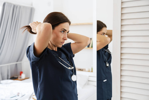 Focused mid woman doctor in medical suit doing hair at home - Foto, Imagen