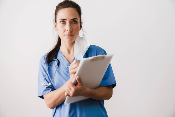 Confident young woman doctor specialist wearing uniform holding notepad isolated over white background, taking notes - Φωτογραφία, εικόνα