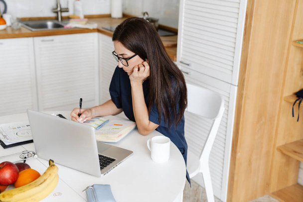 Skupiona kobieta lekarz za pomocą słuchawki podczas pracy z laptopem i papierami w domu, konsultacje online - Zdjęcie, obraz