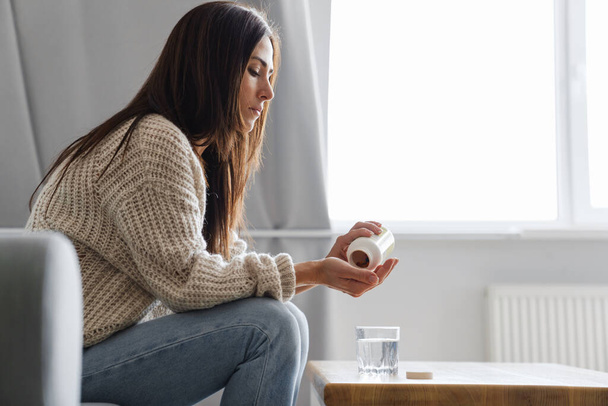 Side view of a young woman holding plastic bottle of pills while sitting on a couch at home with glass of water - Photo, Image