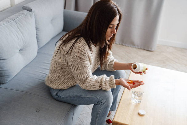 The woman getting ready to take pills while sitting on the couch in front of the table in the room - Photo, image