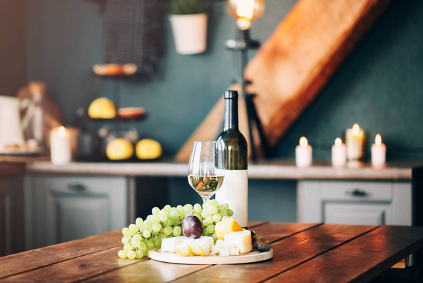Weißweingläser neben köstlichem Käse und grünen Trauben auf Holztisch mit dunkler Küche auf verschwommenem Hintergrund - Foto, Bild