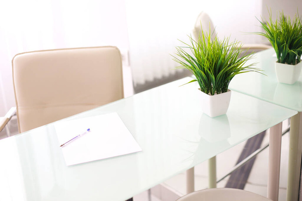 Білий скляний офісний стіл зі світло-бежевим штучним шкіряним стільцем поруч з ним, на фоні дзеркальної стіни. Стільниця прикрашена зеленою рослиною в квітковому горщику і чистим шматочком паперу з ручкою
 - Фото, зображення