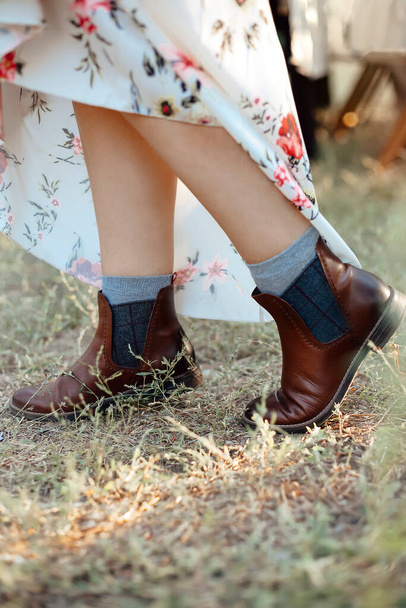 Photo recadrée de jambes féminines regardant de dessous une robe longue et surélevée en cuir naturel mi-saison bottines plates brun foncé, habillées de chaussettes grises, debout à l'extérieur sur de l'herbe sèche d'automne - Photo, image
