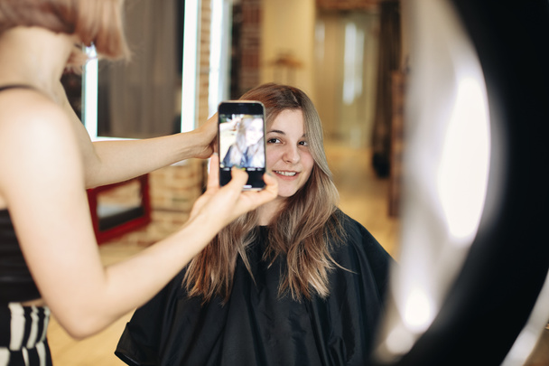 Молодая кавказка парикмахерша делает профессиональное фото клиентов перед яркой лампой в своем портфолио после окончания клиентской прически, используя смартфон. Красота и прическа - Фото, изображение