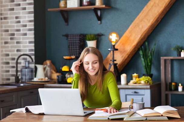 Jeune femme heureuse pigiste assise à table à la maison avec un ordinateur portable, prenant des notes, regardant attentivement dans l'air, pensant à de nouvelles idées, blogueuse à la recherche d'inspiration. Concept freelance - Photo, image