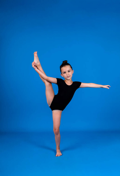 a small gymnast in a black swimsuit does stretching exercises on a blue background with a place for text - Фото, зображення