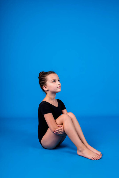 Маленькая гимнастка в черном купальнике сидит на синем фоне с копией пространства - Фото, изображение