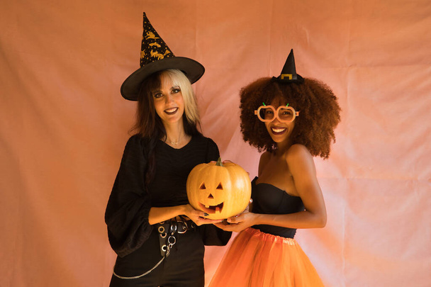 Fiatal latin és spanyol nők boszorkánynak öltözve mosolyognak és egy tököt tartanak rózsaszín alapon Halloweenkor.. - Fotó, kép