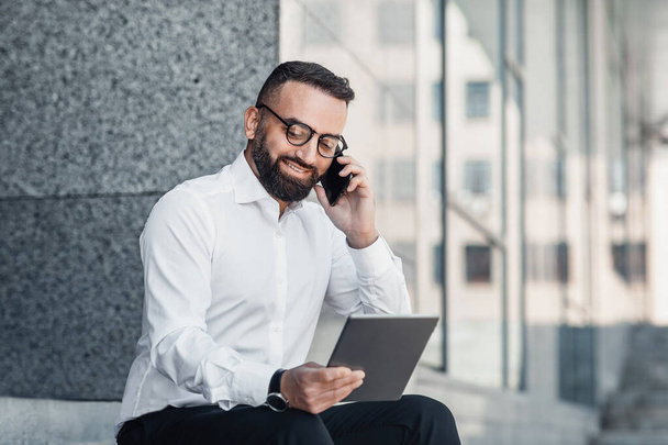 Schöner reifer Mann telefoniert, hält ein digitales Tablet in der Hand und sitzt in der Nähe des Bürozentrums - Foto, Bild