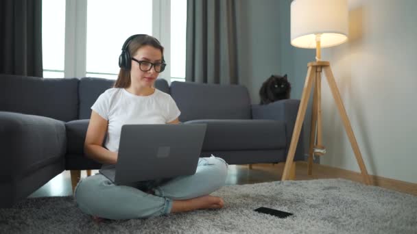 Femme vêtue occasionnellement avec casque est assis sur le tapis avec ordinateur portable et de travailler dans une chambre confortable. Travail à distance en dehors du bureau. - Séquence, vidéo