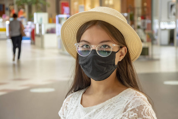 Porträt eines jungen, modischen Mädchens mit Strohhut, 17-20 Jahre alt, Brille und medizinischer Maske aus einem Einkaufszentrum. Ein normaler Kunde im Supermarkt - Foto, Bild