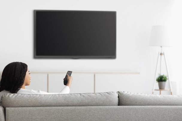 Αφροαμερικανή γυναίκα βλέποντας τηλεόραση δείχνοντας τηλεχειριστήριο σε επίπεδη οθόνη πλάσμα τηλεόραση με λευκή οθόνη, mockup - Φωτογραφία, εικόνα