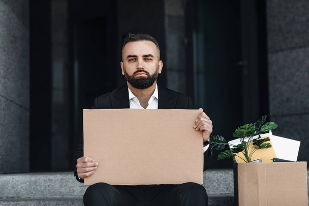 Retrato de cintura hacia arriba del empleado molesto que busca trabajo, sentado afuera y sosteniendo un póster vacío, espacio para copiar - Foto, Imagen