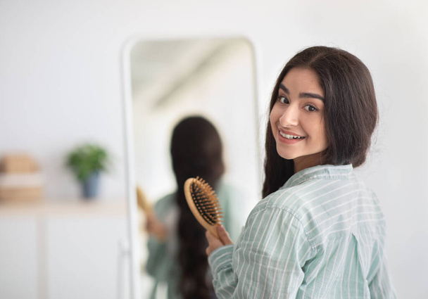 鏡の前に木製のブラシで立って長い美しい髪を持つ魅力的なインドの女性,屋内カメラを見て - 写真・画像