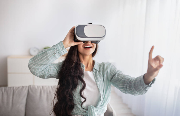 Ευτυχισμένη Ινδή γυναίκα με γυαλιά VR που αλληλεπιδρά με την εικονική πραγματικότητα, εξερευνά τον κυβερνοχώρο, παίζει βιντεοπαιχνίδια στο σπίτι - Φωτογραφία, εικόνα