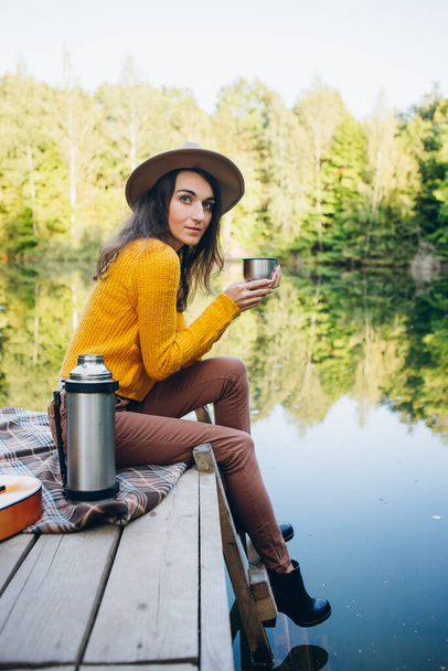 Young woman sits on a bridge on a lake with an autumn landscape and drinks hot tea from a thermos. Toning. - Fotografie, Obrázek