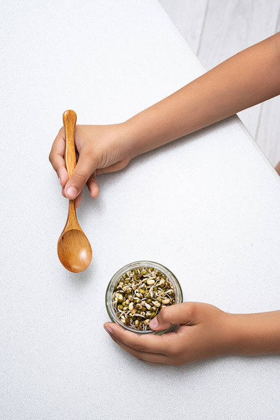 Sprießende Mungbohnen in einem Glas in den Händen eines Kindes. Mungbohnen Zutaten verschiedener asiatischer Gerichte. Veganes oder Rohkost-Ernährungskonzept. - Foto, Bild