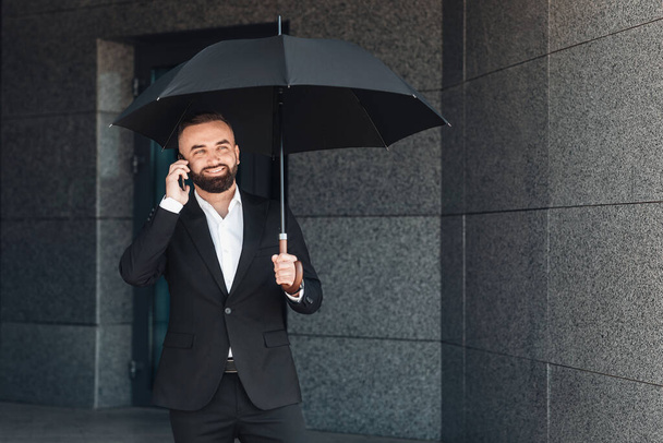 Magabiztos üzletember öltönyben telefonál, miközben fekete esernyőt tart az irodából. - Fotó, kép