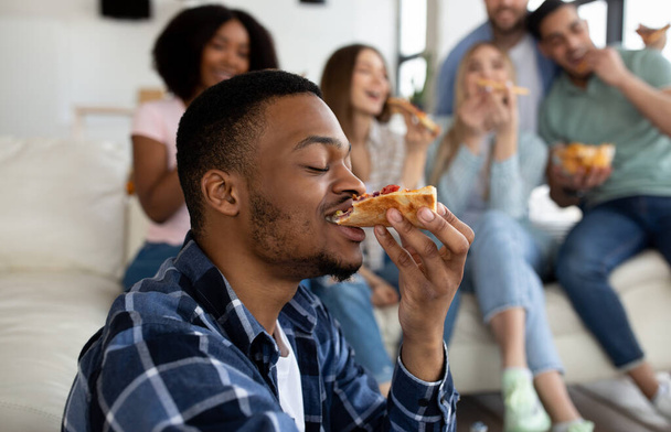 Funky czarny facet jedząc smaczną pizzę ze swoimi wielonarodowymi przyjaciółmi, korzystając towarzyskie spotkanie w domu - Zdjęcie, obraz