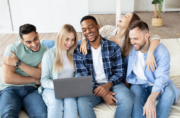Счастливые мультиэтнические друзья, пользующиеся ноутбуком, сидящие на диване, смотрящие кино, имеющие видеозвонки из дома - Фото, изображение