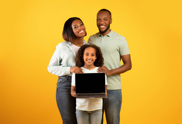 Excited black family of three showing laptop with blank black screen, smiling girl holding gadget with free space - 写真・画像